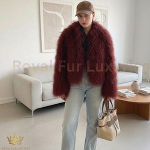 May include: A burgundy-colored, shaggy fur jacket. The jacket is worn with light-wash jeans and a brown baseball cap. A beige and brown handbag is held in the right hand. The background includes a light-colored sofa and a coffee table.