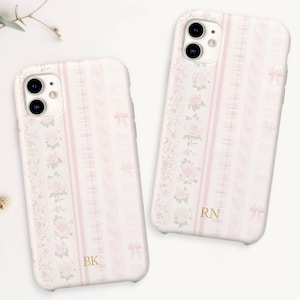 May include: Two white phone cases with a pink floral and ribbon pattern. The cases have gold initials and are decorated with pink roses and bows. A sprig of eucalyptus and small white flowers are in the background.