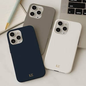 Może przedstawiać: Trzy etui na telefon w kolorze granatowym, szarym i białym, każde ze złotym monogramem i modułem aparatu. Notatnik, długopis i klawiatura laptopa znajdują się w tle. Białe etui ma złote litery "KE".