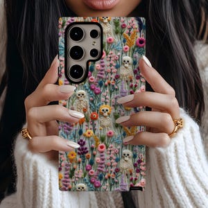 May include: A phone case with a floral design featuring embroidered skulls and flowers. The case is black with a white floral pattern and has a camera cutout.