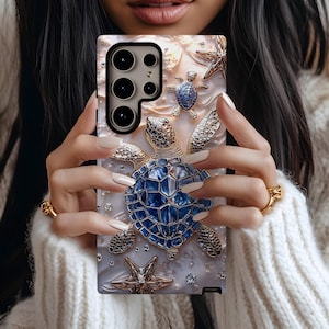 Puede incluir: Una funda de teléfono con un diseño 3D de una tortuga marina azul enjoyada, estrellas de mar y otra vida marina. La funda tiene un fondo nacarado y detalles metálicos. La funda se sostiene frente a un suéter blanco.