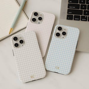 May include: Three phone cases with a gingham pattern in light pink, light blue, and off-white. The cases have a black border and a camera cutout. One case has the initials "KC" and another has "CK". A notebook, pen, and laptop are also in the image.