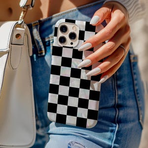 May include: A smartphone case with a black and white checkerboard pattern. The case has a pearlescent finish and a camera cutout. The phone case is held in front of a pair of blue jeans and a white purse.