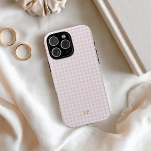May include: A pink and white gingham patterned phone case with a black border. The case has a gold monogram of the letters "AP" and a camera lens. The phone case is on a white fabric surface.