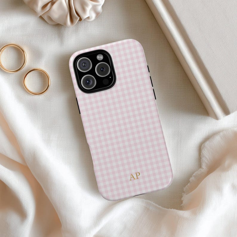 May include: A pink and white gingham patterned phone case with a black border. The case has a gold monogram of the letters "AP" and a camera lens. The phone case is on a white fabric surface.