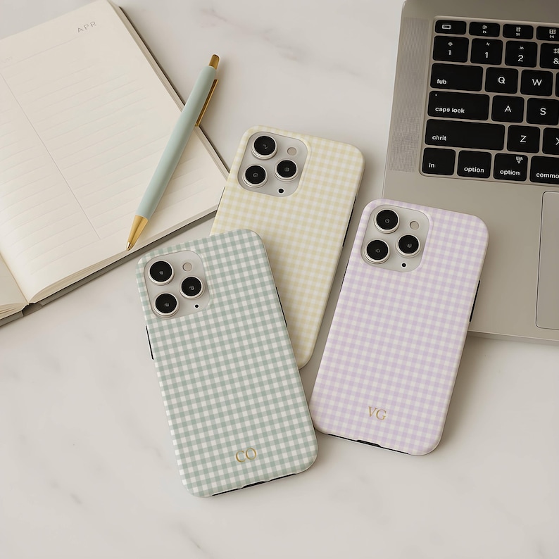 May include: Three phone cases with a gingham pattern in pastel colors: green, yellow, and lavender. The cases are on a white surface next to a laptop, a notebook, and a pen. The green case has the letters "CO", and the lavender case has the letters "VC".