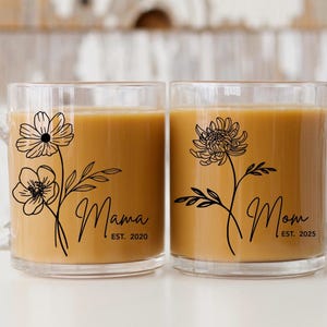 May include: Two clear glass mugs filled with a light brown liquid. Each mug features a black floral design and the words "Mama" and "Mom" in script, with the years "EST. 2020" and "EST. 2025" below. The mugs have clear handles.