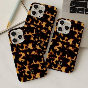 May include: Three phone cases with a tortoiseshell pattern in shades of brown and yellow are displayed. Each case has a camera cutout and a personalized monogram. One case has "ST", and another has "AS". The cases are on a white surface.