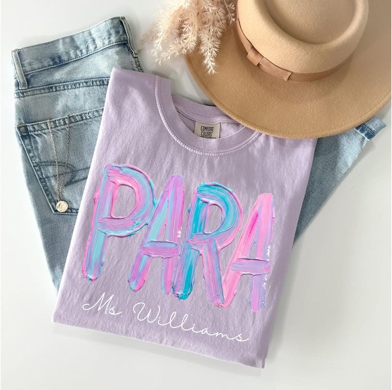 May include: A lilac-colored t-shirt with the word "PARA" in a colorful, brushstroke design. Below the word is the name "Ms. Williams" in a delicate script. The shirt is paired with blue jeans and a tan hat.
