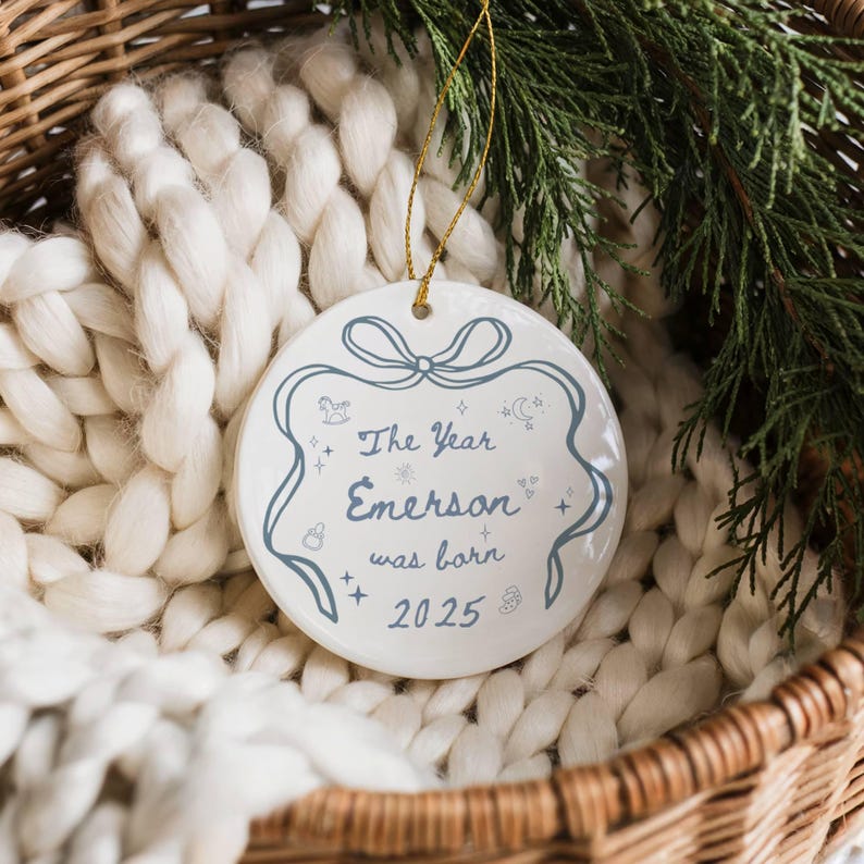 K&ouml;nnte beinhalten: Ein wei&szlig;er Keramik-Weihnachtsschmuck mit goldenem Band, mit dem Text "The Year Emerson was born 2025" in blauer Schrift. Der Schmuck hat einen dekorativen blauen Rand mit Sternen und anderen kleinen Illustrationen.