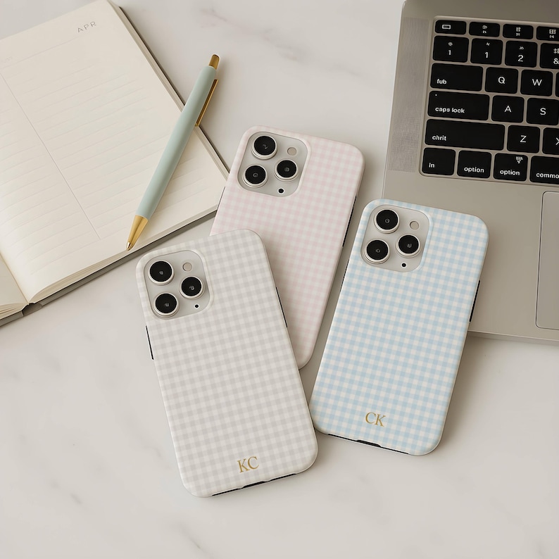 May include: Three phone cases with a gingham pattern in light pink, light blue, and light gray. The cases have a camera cutout and personalized initials. A pen and notebook are in the background.