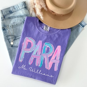 May include: A purple t-shirt with the word "PARA" in a colorful, brushstroke design. The name "Ms. Williams" is written below. The shirt is paired with light-wash jeans and a tan hat.