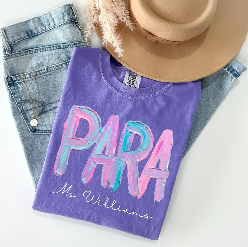 May include: A purple t-shirt with the word "PARA" in a colorful, brushstroke design. The name "Ms. Williams" is written below. The shirt is paired with light-wash jeans and a tan hat.