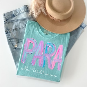 May include: A light blue t-shirt with the word "PARA" in large, textured pink and blue letters. The name "Ms. Williams" is written below. A tan hat and light wash jeans are also in the image.