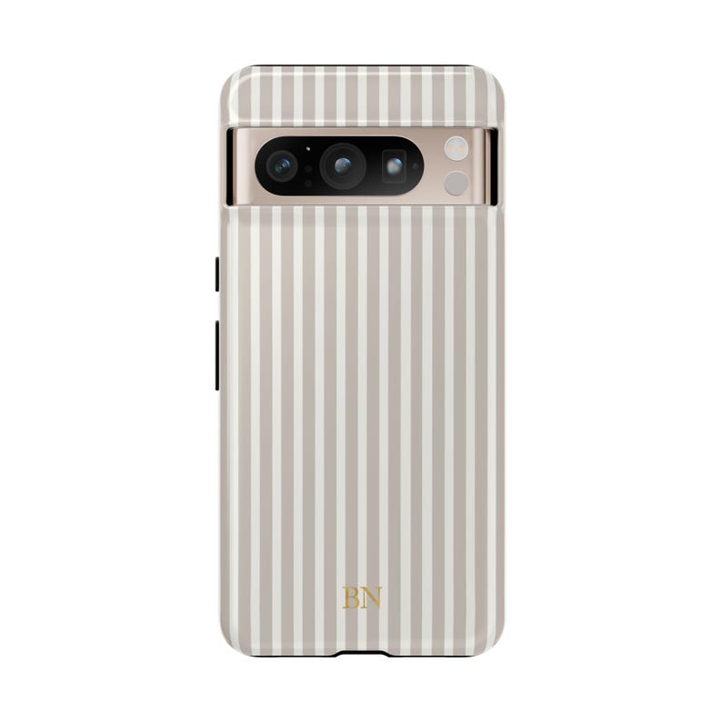 以下が含まれることがあります： ライトグレーと白のストライプ模様の携帯電話ケース。ケースにはカメラの切り抜きがあり、下部には金色の文字「BN」があります。この携帯電話ケースは、特定の電話モデルに合うように設計されています。