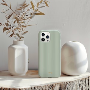 以下が含まれることがあります： 縦縞模様のセージグリーンのiPhoneケース。金色のイニシャル「WR」入り。乾燥した葉が入った白い花瓶と、丸みを帯びた白いセラミックの花瓶が木製の表面に置かれています。