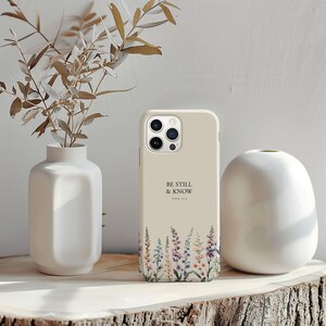 May include: A beige phone case with the text "BE STILL & KNOW" and a floral design at the bottom. The case is displayed on a wooden surface next to a white vase and a decorative plant.
