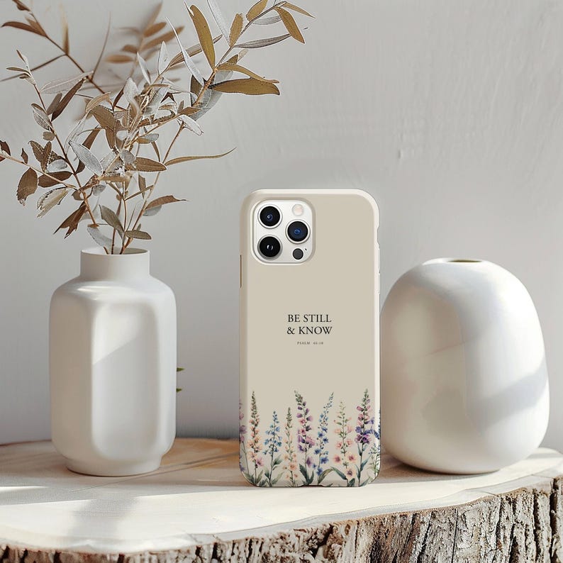 May include: A beige phone case with the text "BE STILL & KNOW" and a floral design at the bottom. The case is displayed on a wooden surface next to a white vase and a decorative plant.