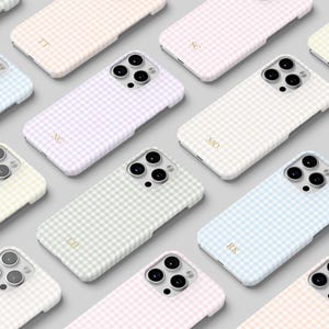 May include: A collection of pastel-colored phone cases arranged on a light gray surface. Each case features a gingham pattern in shades of pink, blue, green, and yellow. Some cases have gold initials. The cases are designed for a smartphone.