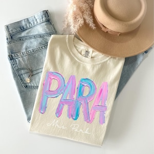 May include: Cream-colored t-shirt with the word "PARA" in large, textured, blue and pink brushstrokes. Below the word is the name "Mrs. Park" in cursive. The shirt is paired with light-wash jeans, a tan hat, and a necklace.