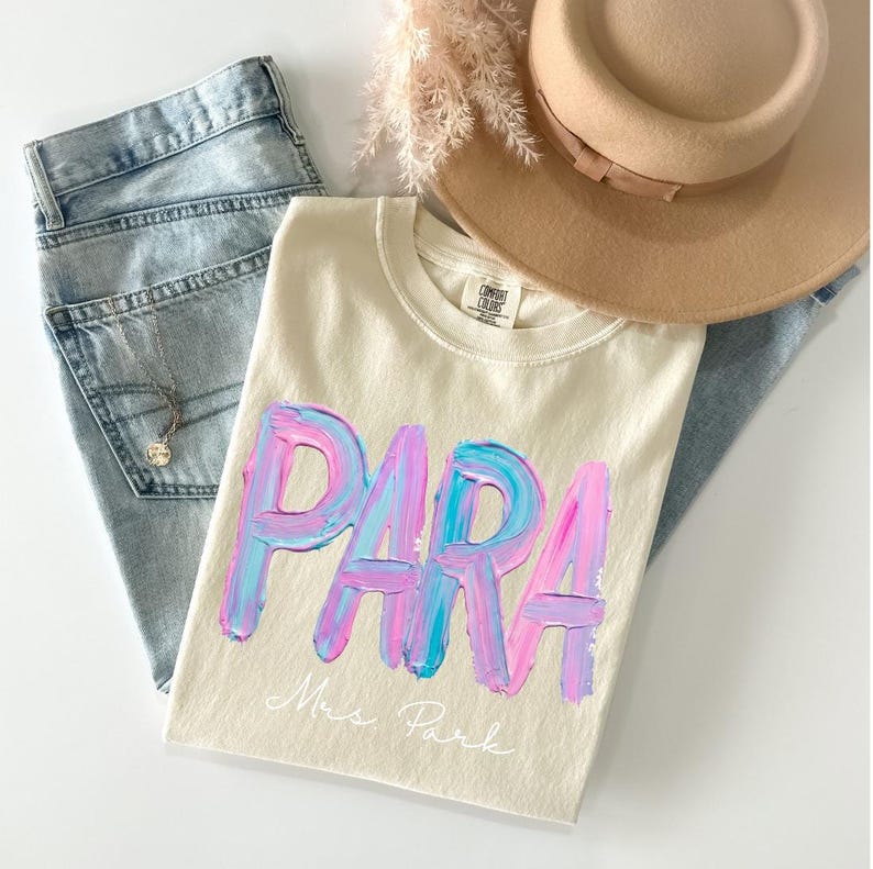 May include: Cream-colored t-shirt with the word "PARA" in large, textured, blue and pink brushstrokes. Below the word is the name "Mrs. Park" in cursive. The shirt is paired with light-wash jeans, a tan hat, and a necklace.