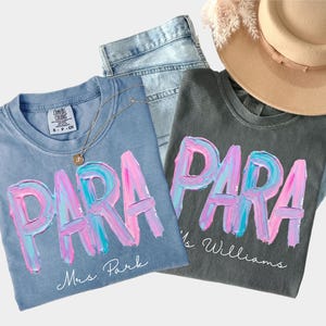 May include: Two t-shirts with the word "PARA" in a colorful, brushstroke design. One is light blue with "Mrs. Park" below, the other is gray with "Ms. Williams". A gold necklace, jeans, and a beige hat are also in the image.