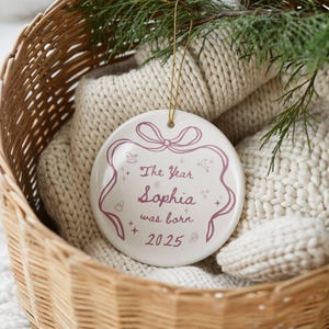 K&ouml;nnte beinhalten: Wei&szlig;er Keramik-Weihnachtsschmuck mit goldenem Faden, mit dem Text "The Year Sophia was born 2025" in Malve. Der Schmuck ist mit einem Band, Sternen und festlichen Symbolen verziert. Er liegt in einem geflochtenen Korb mit einer Strickdecke.