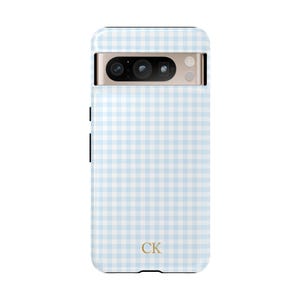 May include: A light blue and white gingham patterned phone case. The case has a camera cutout and the initials "CK" in gold at the bottom. The phone case is designed to fit a specific phone model.