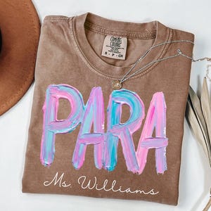 May include: A brown Comfort Colors t-shirt with the word "PARA" in large, textured, pink and blue brushstrokes. Below the word is the name "Ms. Williams" in cursive. A silver necklace with a pendant is draped across the shirt.
