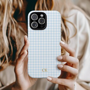 May include: A light blue and white gingham patterned phone case with a gold "CK" monogram. The case has a camera cutout and is held by a person. The case is designed for a smartphone.
