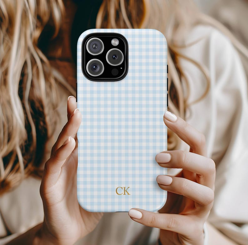 May include: A light blue and white gingham patterned phone case with a gold "CK" monogram. The case has a camera cutout and is held by a person. The case is designed for a smartphone.