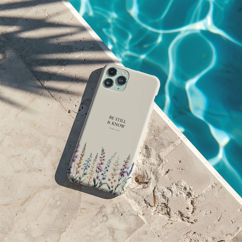 May include: A light beige phone case with a floral design at the bottom and the text "BE STILL & KNOW" in black. The case is lying on a stone surface next to a pool of water.