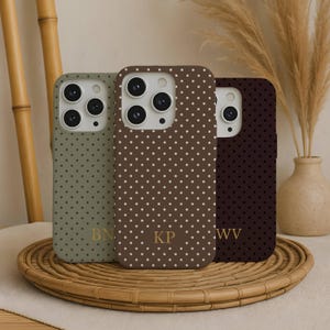 May include: Three phone cases with a polka dot pattern in olive green, brown, and burgundy. Each case has a different set of initials in gold. The cases are displayed on a round, woven tray with a neutral background.