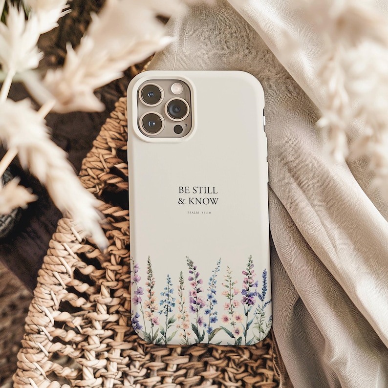 May include: Off-white phone case with the text "BE STILL & KNOW" and Psalm 46:10 printed on it. The case features a floral design with purple, pink, and blue flowers at the bottom. The phone case is resting on a woven basket and a beige cloth.