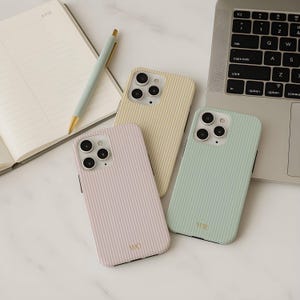 以下が含まれることがあります： パステルカラーの縦縞模様の携帯電話ケース3つ。ケースはライトピンク、ペールイエロー、ミントグリーンです。各ケースにはカメラの切り抜きと金のモノグラムが付いています。背景にはノート、ペン、ラップトップがあります。