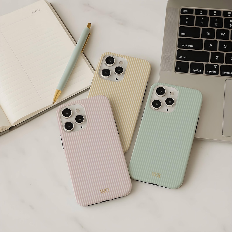 以下が含まれることがあります： パステルカラーの縦縞模様の携帯電話ケース3つ。ケースはライトピンク、ペールイエロー、ミントグリーンです。各ケースにはカメラの切り抜きと金のモノグラムが付いています。背景にはノート、ペン、ラップトップがあります。