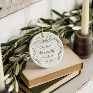 K&ouml;nnte beinhalten: Wei&szlig;er Keramik-Ornament mit goldenem Aufh&auml;nger. Der runde Anh&auml;nger tr&auml;gt den gr&uuml;nen Text "The Year Kennedy was born 2025". Das Design umfasst eine Schleife und weitere dekorative Elemente.