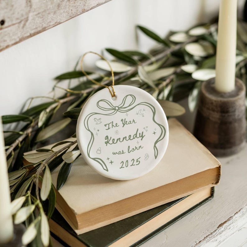 K&ouml;nnte beinhalten: Wei&szlig;er Keramik-Ornament mit goldenem Aufh&auml;nger. Der runde Anh&auml;nger tr&auml;gt den gr&uuml;nen Text "The Year Kennedy was born 2025". Das Design umfasst eine Schleife und weitere dekorative Elemente.