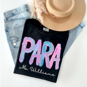 May include: Black t-shirt with the word "PARA" in a colorful, textured font, with "Ms. Williams" below. The shirt is paired with light-wash jeans, a tan hat, and a gold necklace.
