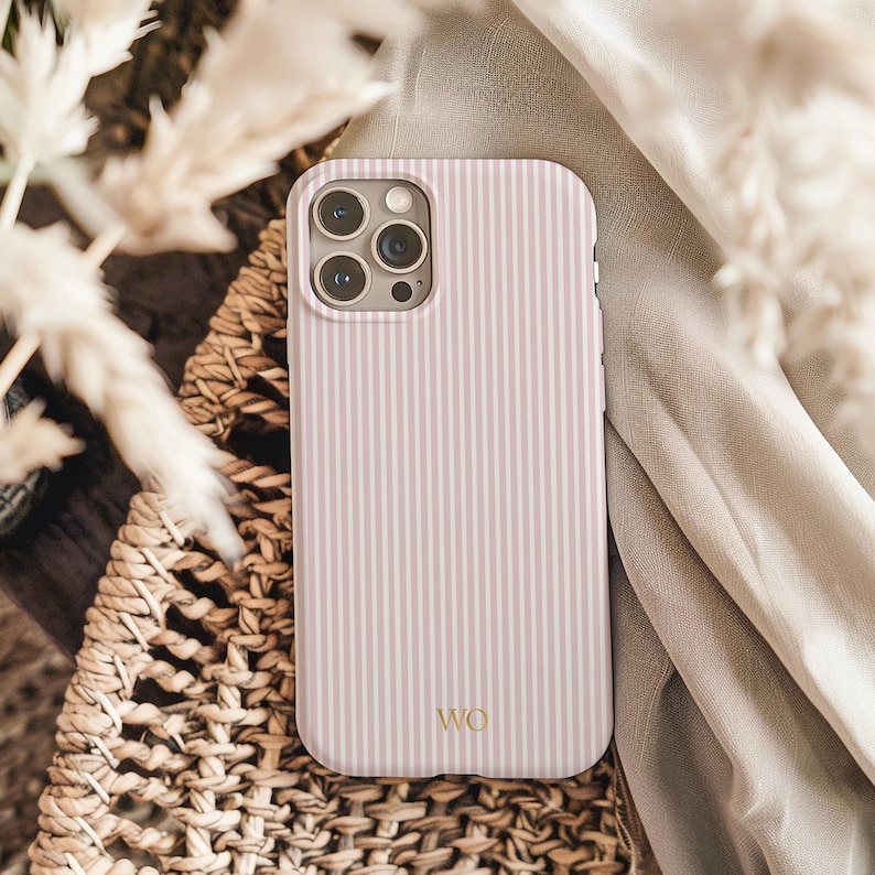 以下が含まれることがあります： ピンクと白のストライプの携帯電話ケース。金色の文字で「WO」のイニシャルが入っています。ケースは、織りのバスケットとベージュの生地の上に表示されています。ケースにはカメラの切り抜きがあります。