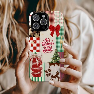 May include: A colorful phone case with a patchwork design. The case features a red bow, a Christmas tree, and the text "He Restores my Soul." The design includes red and white checkered patterns, stars, and a cowhide print.