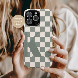 May include: A white and grey checkered phone case with a green letter K in the centre. The case is designed for a phone with a square camera lens.