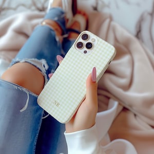 May include: A light yellow and white gingham pattern phone case with three camera lenses. The case has the initials "NR" in the bottom corner. The phone case is held in a person's hand.