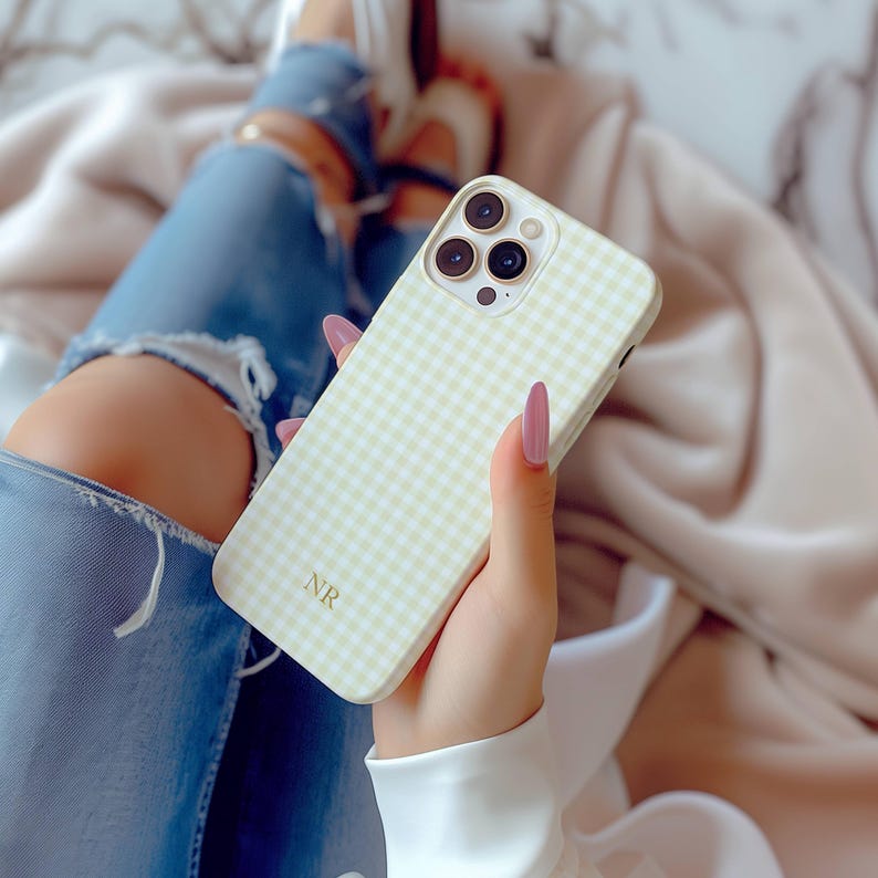 May include: A light yellow and white gingham pattern phone case with three camera lenses. The case has the initials "NR" in the bottom corner. The phone case is held in a person's hand.