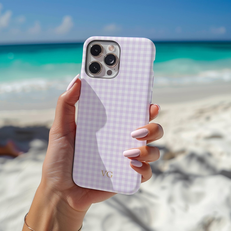 May include: A light purple and white gingham patterned phone case. The case has a gold "VG" monogram. The phone case is held up against a beach background with turquoise water and white sand.