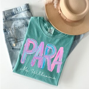 May include: A teal t-shirt with the word "PARA" in pink and blue brushstrokes, with "Ms. Williams" in script below. The shirt is paired with light wash jeans, a tan hat, and a necklace.