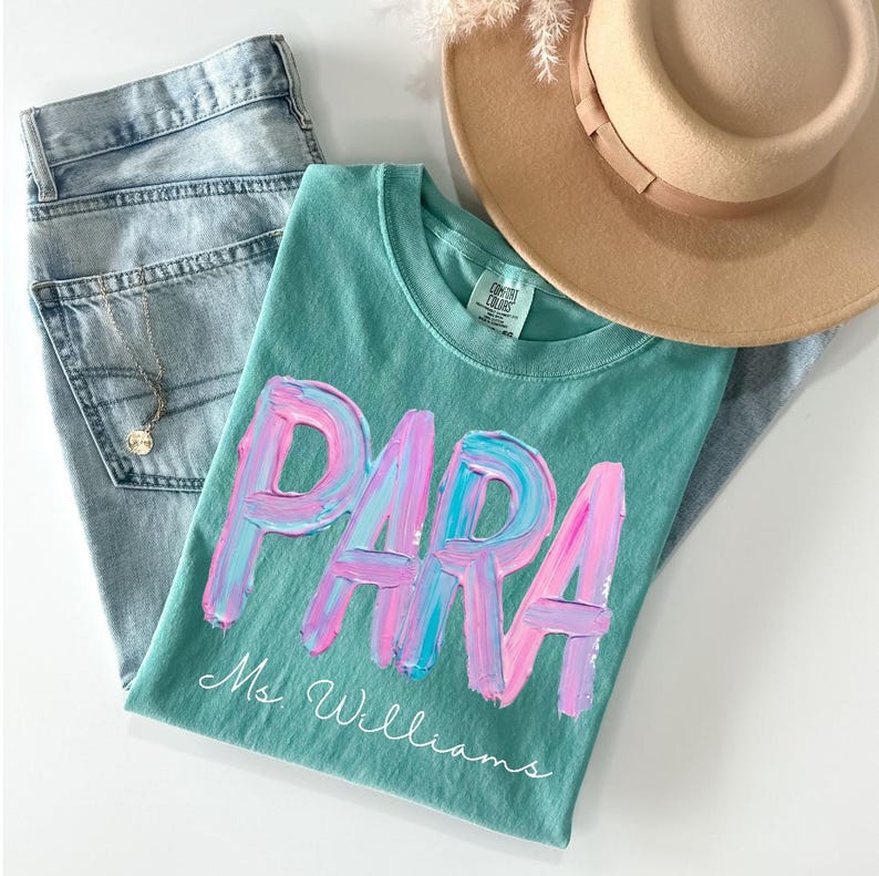May include: A teal t-shirt with the word "PARA" in pink and blue brushstrokes, with "Ms. Williams" in script below. The shirt is paired with light wash jeans, a tan hat, and a necklace.