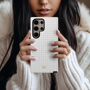 May include: A white and gray gingham patterned phone case with a black camera module. The case has gold initials "KC" at the bottom. The phone case is held by a person wearing a white sweater.