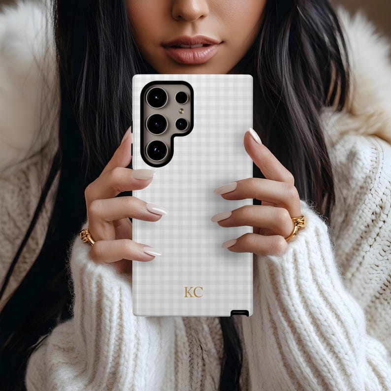 May include: A white and gray gingham patterned phone case with a black camera module. The case has gold initials "KC" at the bottom. The phone case is held by a person wearing a white sweater.