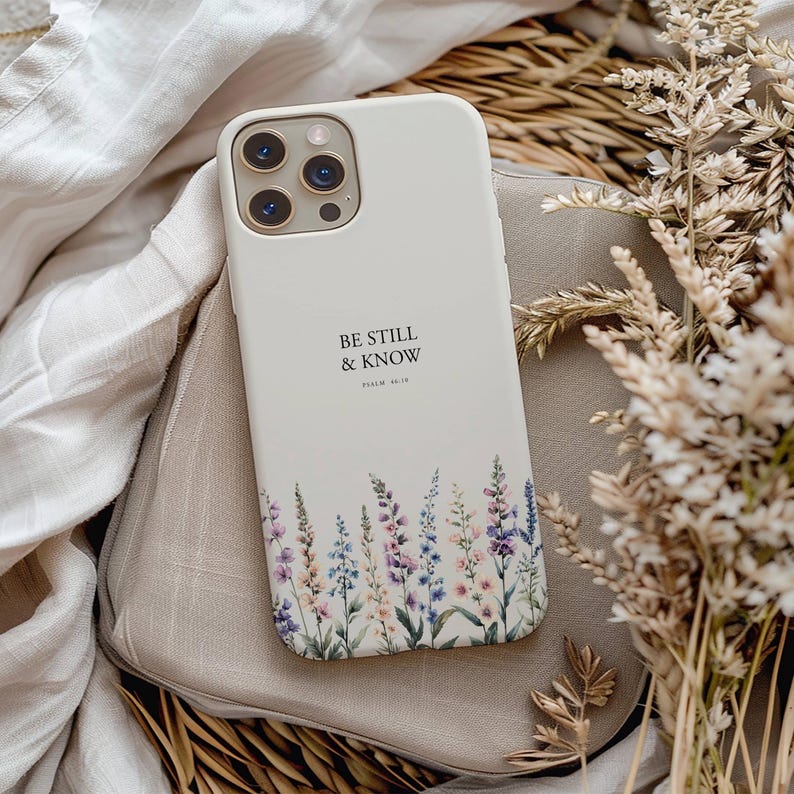 May include: A white iPhone case with the text "BE STILL & KNOW" and Psalm 46:10 printed on it. The case features a floral design with purple, blue, and pink flowers at the bottom. The phone case is resting on a beige surface.